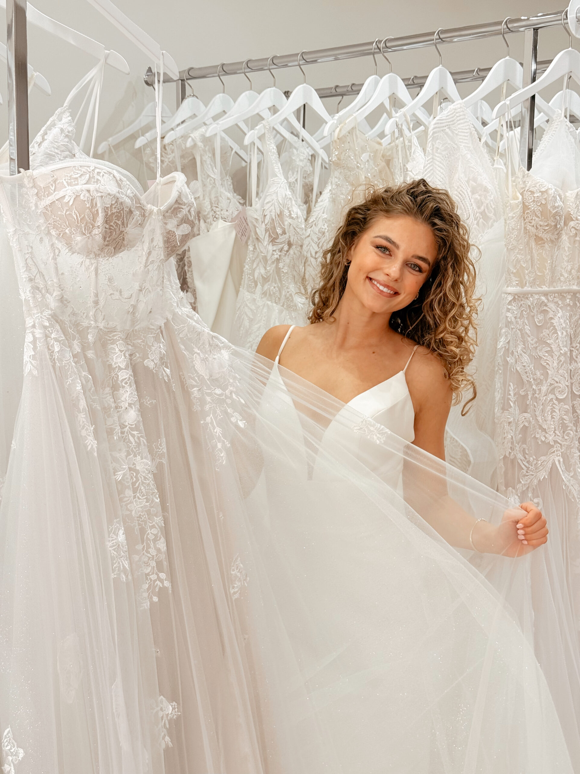 a woman with long, curly blonde hair smiling in a bridal boutique. She is positioned behind several white and ivory wedding dresses hanging on a rack.