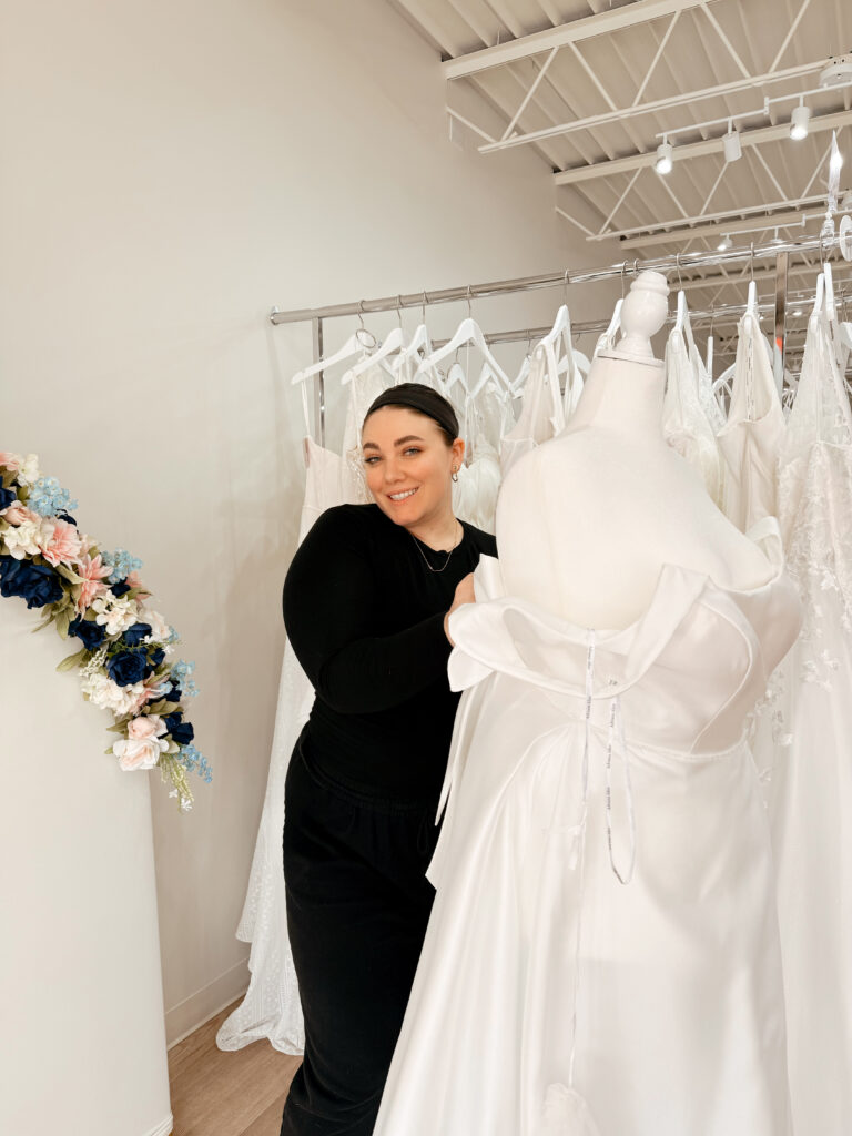 A stylist in black attire smiles while adjusting a white gown on a mannequin, set against a background of dress racks and a floral wall installation.