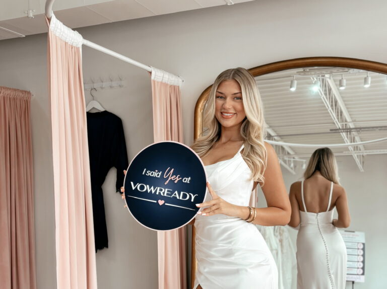 happy blonde woman wearing a white bridal gown holding a blue signage.