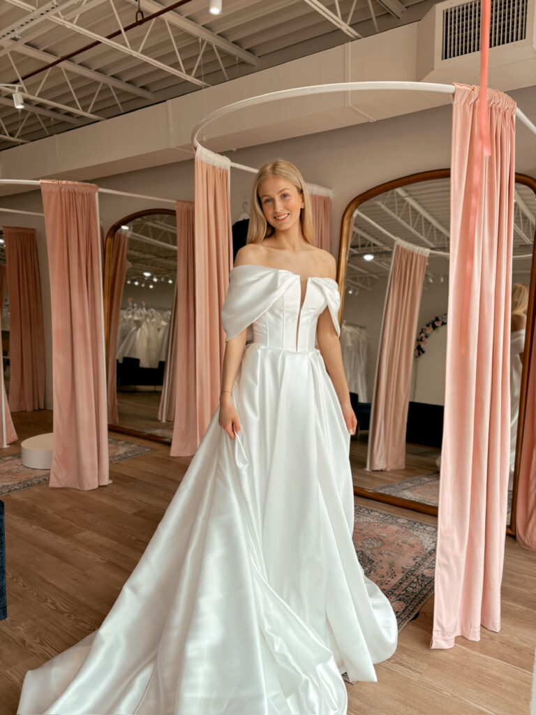 Bride wearing an off-the-shoulder satin A-line wedding dress with a plunging neckline during a bridal appointment at Savvy Bridal.