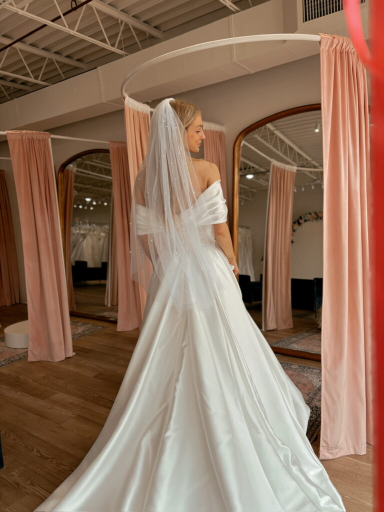 A bride models an elegant off-the-shoulder satin A-line wedding dress featuring a structured bodice and flowing skirt in a private bridal suite at Savvy Bridal.