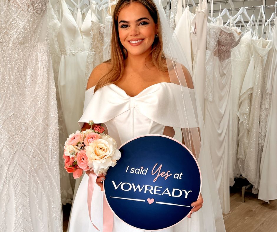 Bride wearing an off-the-shoulder wedding dress and holding a bouquet with a Vow Ready sign inside the bridal boutique.