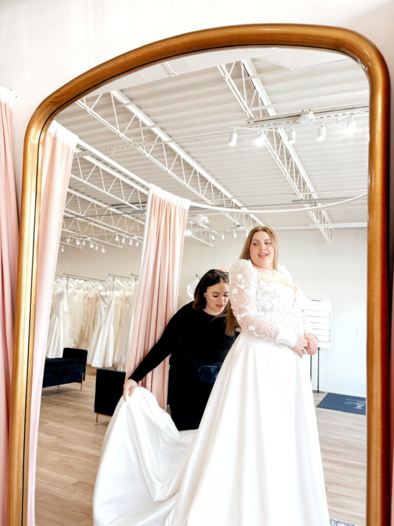Bridal stylist helping a bride with her wedding dress in front of a mirror.