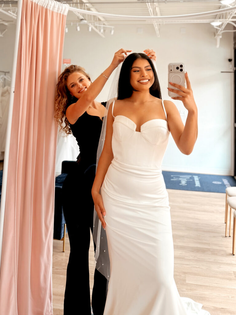Bridal stylist adjusting a bride’s veil as she takes a mirror selfie in her wedding dress.